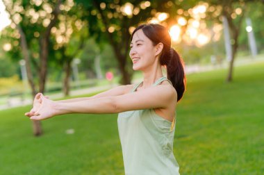 Kadın koşucu. Açık havada koşup eğlenmeden önce yeşil spor kıyafetli sağlıklı bir Asyalı genç kadın parkta kaslarını esnetiyor. Kamu parkındaki spor koşucusu kız. Refah konsept demek.