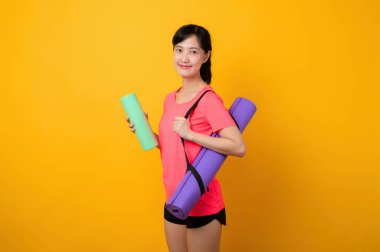 Asyalı genç spor fitness kadını portresi. Pembe spor kıyafetleri ve yoga minderi giymiş mutlu bir gülümseme. Sarı stüdyo arka planında egzersiz yapıyor. Sağlıklı sağlık yaşam tarzı kavramı.