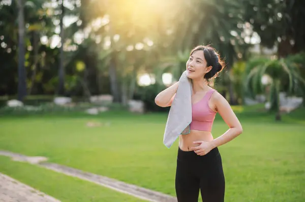 Happy asian 30s woman in pink sportswear wipes her cheek after sunset workout in nature. Captures beauty of healthy lifestyle and perfect for project promoting fitness, health, or motivation.