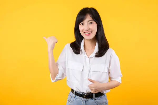 ハッピーアジアの若い女性は白いシャツを着て 黄色の背景に隔離されたコピースペースに親指を指差すデニムのジーンズ植物 最もよい選択を選ぶのを助け プロダクト概念を推薦して下さい — ストック写真
