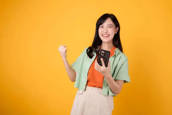 Radiate confidence and empowerment of Asian woman 30s wearing orange and green shirt. confidently raises fist while using smartphone, showing determination and digital prowess on yellow background.
