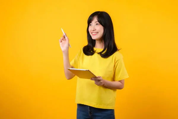 亚洲的年轻女性穿着黄色的T恤和牛仔裤 在使用数码平板电脑时露出快乐的笑容 表现出深思熟虑的表情和创意 教育技术创新思维概念 — 图库照片