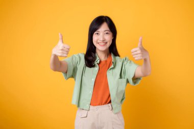 30 'lu yaşlarda, turuncu gömlekli, yeşil kazaklı, zarif bir Asyalı kadın. Sarı arka planda yaptığı jest, onaylama kavramının somut bir örneği..