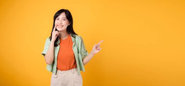 Büyüleyici 30 'lu yaşlarda Asyalı bir kadının diş sağlığı, dişlerine turuncu tişört ve yeşil kazak izi takması, parlak sarı arka planda izole edilmesi, diş sağlığı hizmetlerinin önemini vurgulaması,.