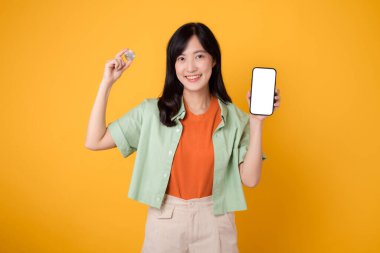 Finans dünyasının geleceğini, turuncu tişört ve yeşil kazak giyen, sarı arka planda akıllı telefon ekranı ve kripto para gösteren büyüleyici Asyalı 30 'lu yaşlarda genç bir kadınla kucaklayın..