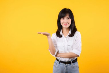 Beyaz gömlek ve kot pantolon giyen mutlu Asyalı genç kadın sarı arka planda izole edilmiş ürünler için fotokopi alanı olan açık avuç içini gösteriyor ya da gösteriyor. Mevcut ürün reklamcılığı konsepti.