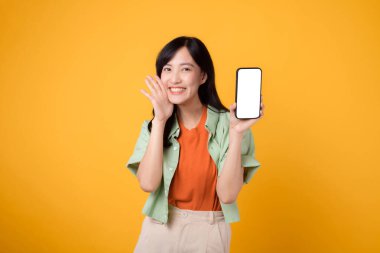 30 yaşlarında, turuncu tişörtlü ve yeşil kazaklı genç Asyalı bir kadın sarı stüdyo arka planında bağıran bir ağızla akıllı telefon ekranı sergiliyor. Yeni mobil aygıt ve çevrimiçi alışveriş konsepti.