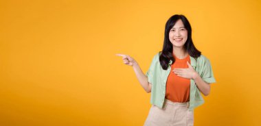 Genç Asyalı 30 'lu yaşlarda, turuncu gömlekli, yeşil tişörtlü, enerjik bir şekilde bedava fotokopi için parmaklarını gösteren ve göğsünü sarı arka planda tutan bir kadın. indirimli alışveriş tanıtım konsepti.