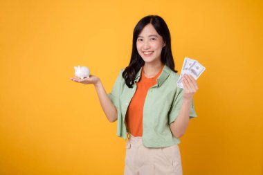 30 yaşlarında, turuncu gömlekli ve yeşil kazaklı genç bir Asyalı kadın. Sarı arka planda dolar ve kumbarayı gösteriyor. Finansal para kavramı.