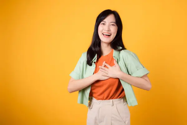 healthcare and wellness with an Asian young woman 30s, wearing a green shirt. She showcases a hand gesture on her chest against a yellow background, embodying the concept of body wellness.