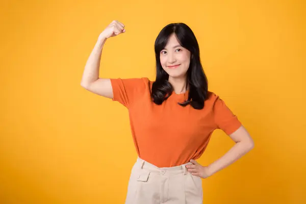 Embrace confidence and feminist empowerment as a young Asian woman 30s raises a fist up hand sign gesture, wearing an orange shirt on yellow background. Empowerment and feminism concept.