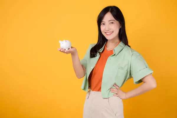 financial money with cheerful young Asian woman 30s, donning orange shirt and green jumper, displaying piggy bank while striking akimbo gesture on yellow background. Financial money concept.