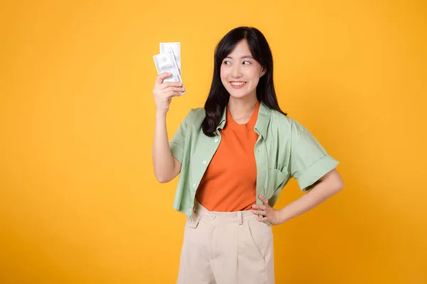 financial money with cheerful young 30s Asian woman, donning orange shirt and green jumper, displaying dollar currency while striking an akimbo gesture on yellow background. Financial money concept.