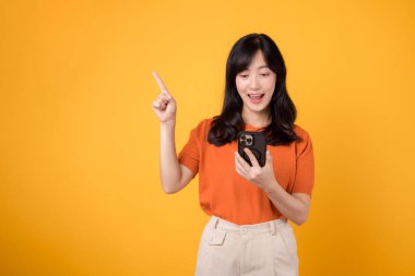 30 'lu yaşlarda genç bir Asyalı kadınla mutluluğu deneyin, turuncu bir gömlek giyiyor, sarı arka planda akıllı telefon kullanırken bedava fotokopi alanını işaret ediyor. yeni uygulama kavramı.