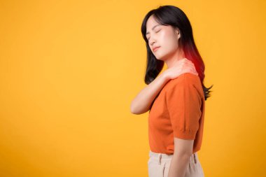 30 'lu yaşlarda, turuncu gömlek giyen Asyalı bir kadın sarı arka planda ağrılı omzunu tutuyor. Boyun ağrısı terapisi tıbbi ofis sendromu konsepti.