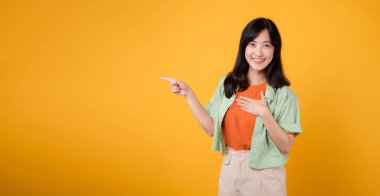 Turuncu tişörte yeşil giyen 30 'lu yaşlardaki genç Asyalı kadın, sarı arka planda göğsünü kapalı tutarken, parmaklarını bedava fotokopi için enerjik bir şekilde işaret ediyor. indirimli alışveriş tanıtım konsepti.