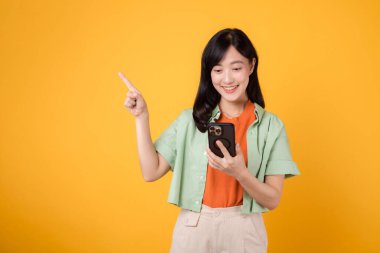 Portakal rengi tişört giyen, sarı arka planda akıllı telefonu kapalı tutarak boş alana parmak işareti yapan büyüleyici 30 'lu yaşlardaki Asyalı bir kadının mobil uygulamaları. uygulama akıllı telefon kavramı.