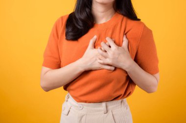 Otuzlu yaşlarda, turuncu tişört giyen, sarı arka planda göğsüne dokunan Asyalı bir kadını yakından çek. Kalp krizi, göğüs ağrısı sağlık konsepti.