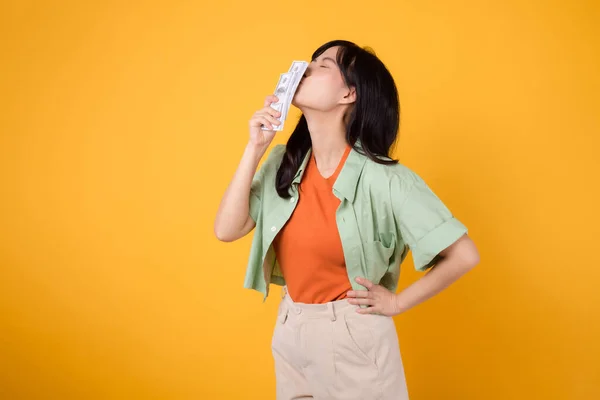 financial money with cheerful young 30s Asian woman, donning orange shirt and green jumper, kissing dollar currency while striking an akimbo gesture on yellow background. Financial money concept.