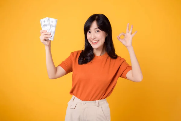 ポジティブな若いアジアの女性 30代 現金ドルを指す オーケーハンドサイン 活気に満ちた黄色の背景に立って — ストック写真
