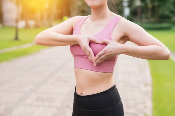 Woman jogger. asian young female happy smile wearing pink sportswear showing heart hands gesture on chest before running in public park. Healthcare wellness concept.
