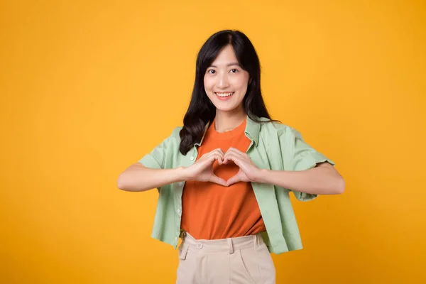 healthcare and wellness as an Asian young woman in her 30s, wearing a green shirt, showcases a heart hand gesture on her chest. Feel the essence of body wellness against a vibrant yellow background.