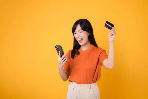 三十多岁的快乐的亚洲女人穿着橙色衬衫 用智能手机 带着黄色背景的信用卡 方便的网上购物 — 图库照片