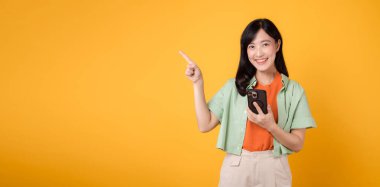 Gezici uygulamalar, turuncu tişört giyen 30 'lu yaşlardaki Asyalı, neşeli genç bir kadının sarı stüdyo arka planında izole edilmiş el hareketleriyle akıllı telefonu gösteriyor. uygulama akıllı telefon kavramı.