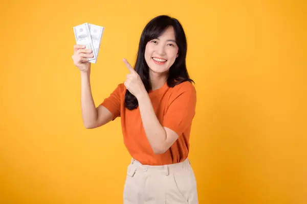 陽気な若いアジアの女性30代 活気に満ちた黄色い背景に立って現金のドルを指して — ストック写真