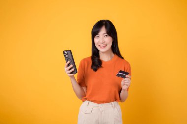 30 'larında turuncu tişörtlü, akıllı telefon kullanan ve sarı arka planda kredi kartı tutan heyecanlı bir Asyalı kadın. İnternetten alışveriş eğlenceli..