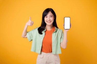 30 'lu yaşlarda, turuncu tişörtlü ve yeşil kazaklı genç Asyalı bir kadın sarı stüdyo arka planında akıllı telefon ekranı gösteriyor. Yeni mobil uygulama kavramı.