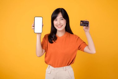 30 'lu yaşlardaki neşeli Asyalı kadın, sarı arka planda akıllı telefon ve kredi kartı sergiliyor. Güvenli çevrimiçi ödeme alışverişi.