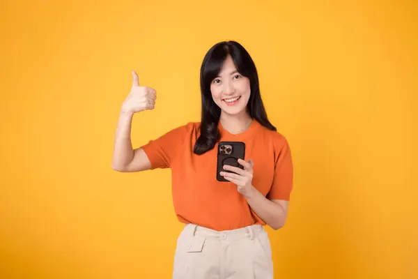 Uśmiechnięta Azjatka Trzydziestce Ubrana Pomarańczową Koszulę Używająca Smartfona Kciukiem Góry — Zdjęcie stockowe