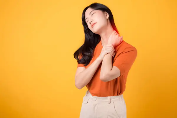 30代の若いアジア人女性は オレンジ色のシャツを着て 黄色の背景に痛みを伴う首を抱えています オフィスシンドロームヘルスケアコンセプト — ストック写真