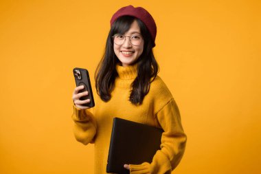 Parlak sarı bir ayarı olan, sarı kazak ve kırmızı bereli genç Asyalı kadın laptopunu ve akıllı telefon görevlerini başarıyla yerine getiriyor..