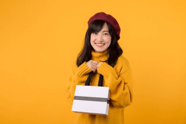 30 'lu yaşlarda genç bir Asyalı kadın, elinde bir alışveriş çantası, sarı bir kazak ve kırmızı bereyle, sarı bir arka planda moda anlayışını sergiliyor..