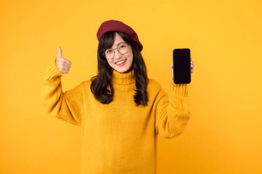 Akıllı telefonlu etnik bir kadın, kırmızı bereli ve gözlüklü, son moda sarı bir ortamda baş parmak hareketi yapıyor..