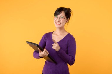 Mor gömlek ve gözlük takan kendine güvenen genç bir kadın, stüdyo portresindeki başarıyı ve profesyonelliği simgeleyen canlı sarı bir arka plana karşı dizüstü bilgisayar tutuyor..