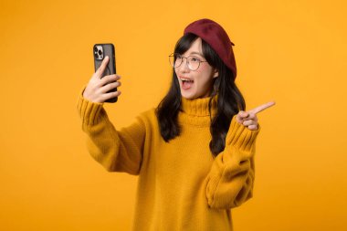 Genç, güzel Asyalı mutlu bir gülümseme takan, sarı gömlekli, kırmızı şapkalı, bedava fotokopi alanına işaret eden ve sarı arka planda akıllı telefon kullanan bir portre. yeni uygulama