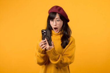 Şaşırmış yüzlü, kırmızı bereli ve sarı kazaklı genç bir kadın, akıllı telefonunu parlak sarı bir arka plana karşı kullanıyor..