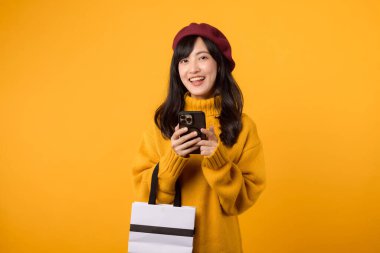 30 'lu yaşlardaki neşeli Asyalı kadın, elinde bir alışveriş çantası ve akıllı telefon, sarı bir kazak ve kırmızı bereyle şık moda anlayışını sergiliyor, alışveriş çılgınlığını kutluyor..
