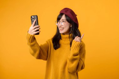 Kırmızı bereli ve sarı kazaklı şık bir kadın akıllı telefonuyla poz veriyor. Anı canlı sarı bir arka planda yakalıyor..