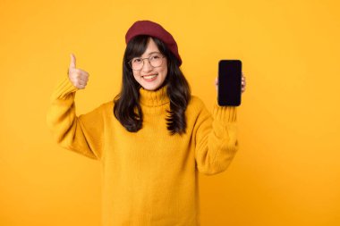 Heyecanlı genç Asyalı kadın sarı kazak kırmızı bere giyiyor ve gözlük takıyor cep telefonu ekranını gösteriyor, parmakları havada, akıllı telefon uygulaması öneriyor, sarı arka planda duruyor.