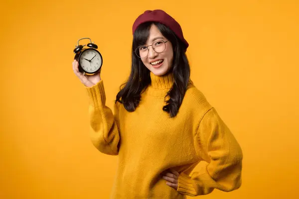 紧挨着生机勃勃的黄色闹钟 一个穿着时髦的黄色毛衣和红色贝雷帽的快乐的年轻亚洲女人 在黄色的背景下流露出一种守时典雅的感觉 — 图库照片