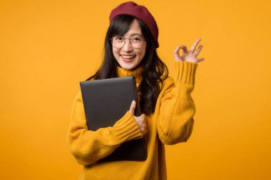 Neşeli sarı ayarı olan, sarı süveter ve kırmızı bereli genç Asyalı bir kadın, dizüstü bilgisayarıyla el hareketi yapıyor..