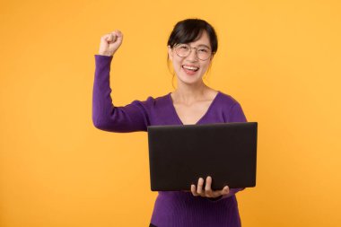 Mor gömlek ve gözlük takan kendine güvenen genç bir kadın dizüstü bilgisayarını ve yumruğunu kaldırıp sarı arka planda başarıyı ve profesyonelliği resmediyor..