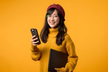 Sarı bir süveter ve kırmızı bereyle, genç Asyalı kadın dizüstü bilgisayarını ve akıllı telefonunu ustalıkla yönetiyor, hepsi de neşeli sarı arka plana karşı..