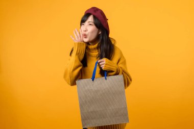 30 'lu yaşlarda, sarı kazaklı ve kırmızı bereli genç bir Asyalı kadın, canlı sarı bir arka plana karşı alışveriş çılgınlığında ve serbest alana jest yaparken zevk alıyor..