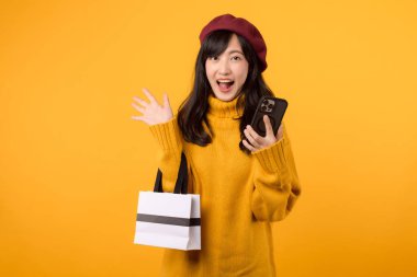 30 'lu yaşlarda genç Asyalı bir kadın, elinde bir alışveriş çantası ve akıllı telefon, sarı süveteri ve kırmızı beresiyle perakende terapisini kutluyor..