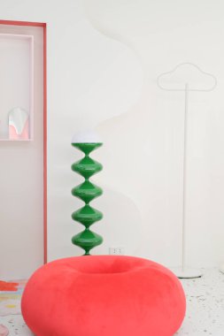 Pembe yuvarlak sandalye, yeşil dekoratif lamba ve minimalist beyaz önlük standı içeren canlı bir iç mekan, neşeli bir atmosfer yaratır.